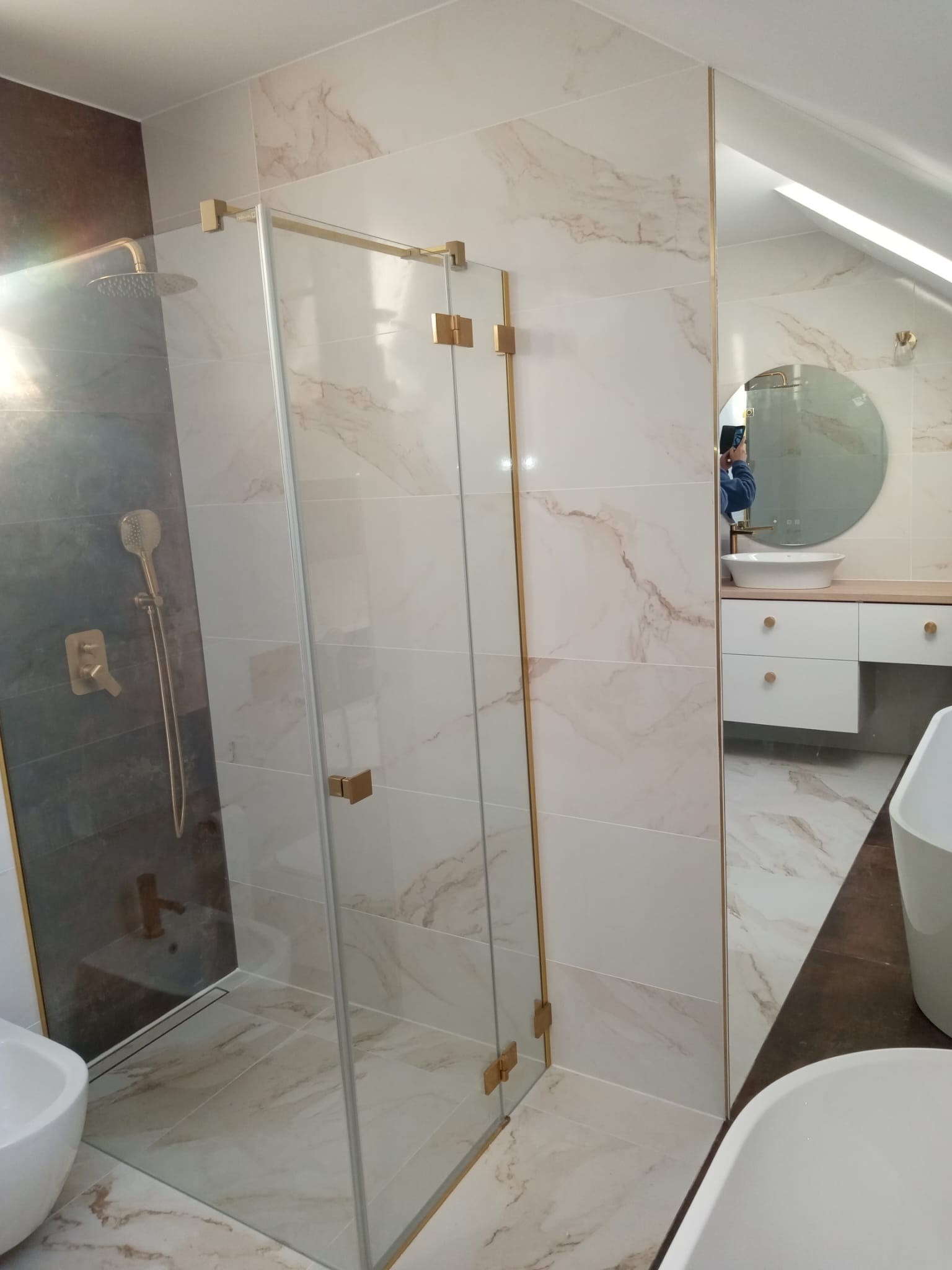 Wykończona łazienka z kabiną prysznicową ze złotymi okuciami, marmurkowymi płytkami na ścianach i podłodze, okrągłym lustrem z podświetleniem LED oraz białą umywalką nablatową i wanną.