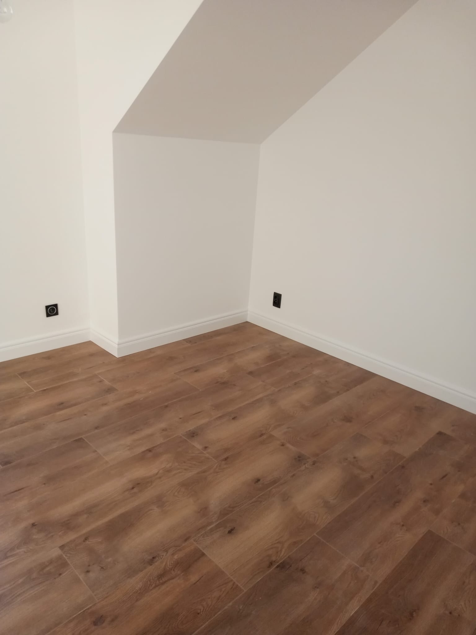 Poddasze po remoncie: brązowe panele podłogowe imitujące drewno, białe ściany z listwami przypodłogowymi i gniazdkami elektrycznymi.