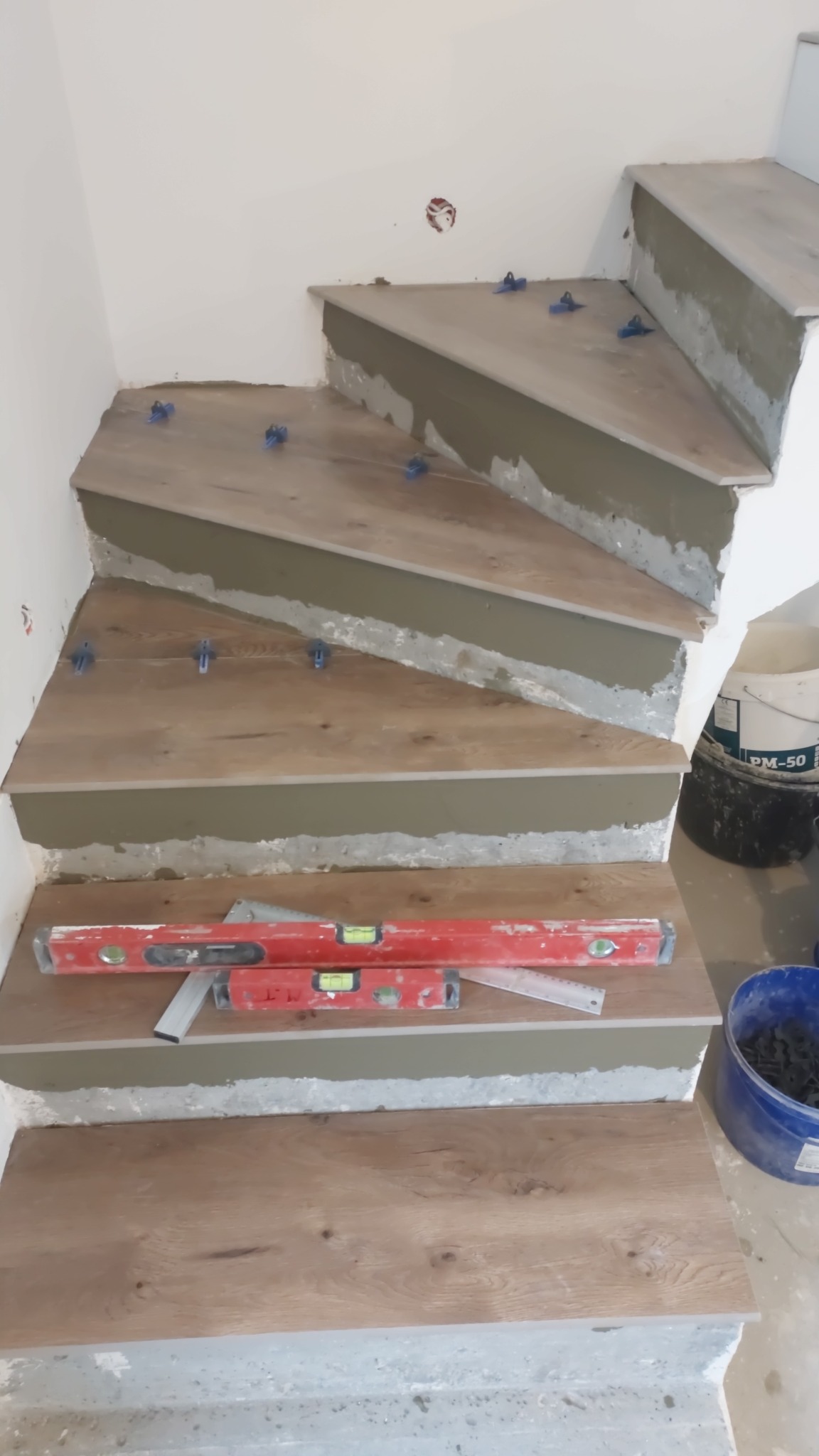 Montaż płytek imitujących drewno na schodach, widoczne poziomice, niebieskie krzyżyki dystansowe i wiadro z zaprawą.