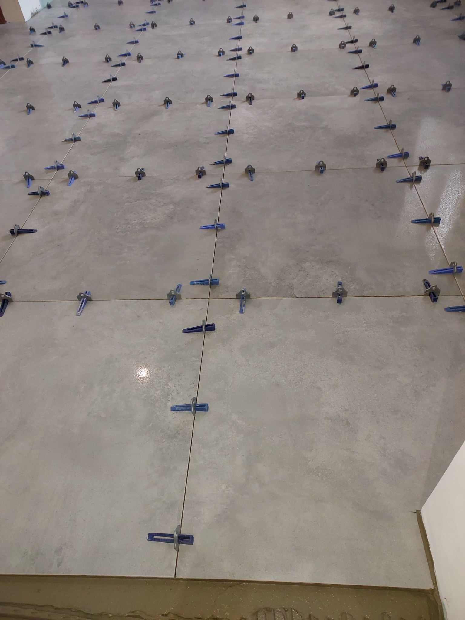 Szare płytki podłogowe w trakcie układania, z użyciem niebieskich klipsów poziomujących, widoczne fugi i zaprawa.