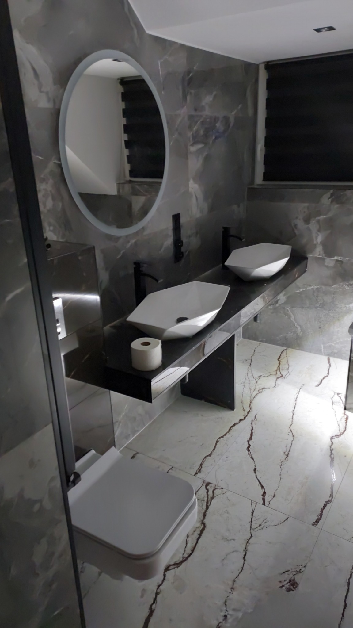 Nowoczesna łazienka z dwiema geometrycznymi umywalkami na czarnym blacie, okrągłe lustro z podświetleniem, marmurowe płytki na ścianach i podłodze z wyrazistym wzorem żyłkowania, wisząca toaleta.