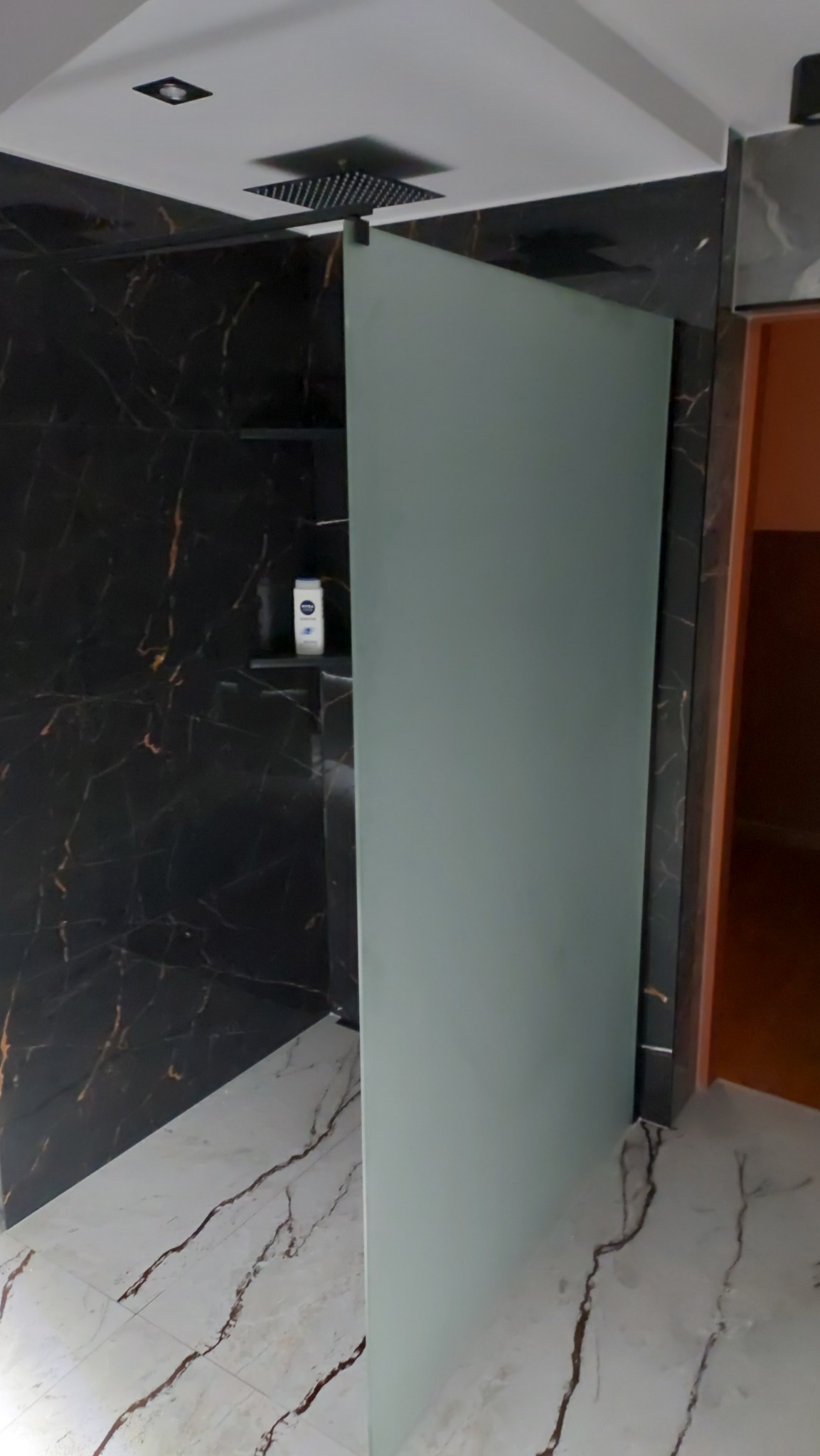 Nowoczesna kabina prysznicowa z matową szybą, czarnymi płytkami imitującymi marmur z złotymi żyłami, biała podłoga z marmurowym wzorem, czarna deszczownica zamontowana w suficie i kosmetyk na półce...