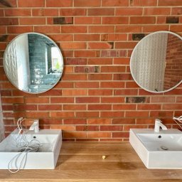 Craftbud - Dwie białe, prostokątne umywalki z bateriami na drewnianym blacie, zamontowane na tle ściany z czerwonej cegły i białych płytek 3D, z okrągłymi lustrami nad nimi, w trakcie montażu instalacji.