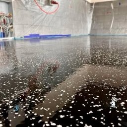 Craftbud - Błyszcząca, ciemna podłoga z białymi drobinkami, świeżo wylana, w pomieszczeniu zabezpieczonym folią malarską, widoczne rury grzewcze przy ścianie.