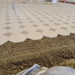 Układanie podłogi z płytek w kształcie ośmiokątów i kwadratów na podsypce piaskowo-cementowej, widoczna linia poziomowania i akcesoria do układania płytek w tle.