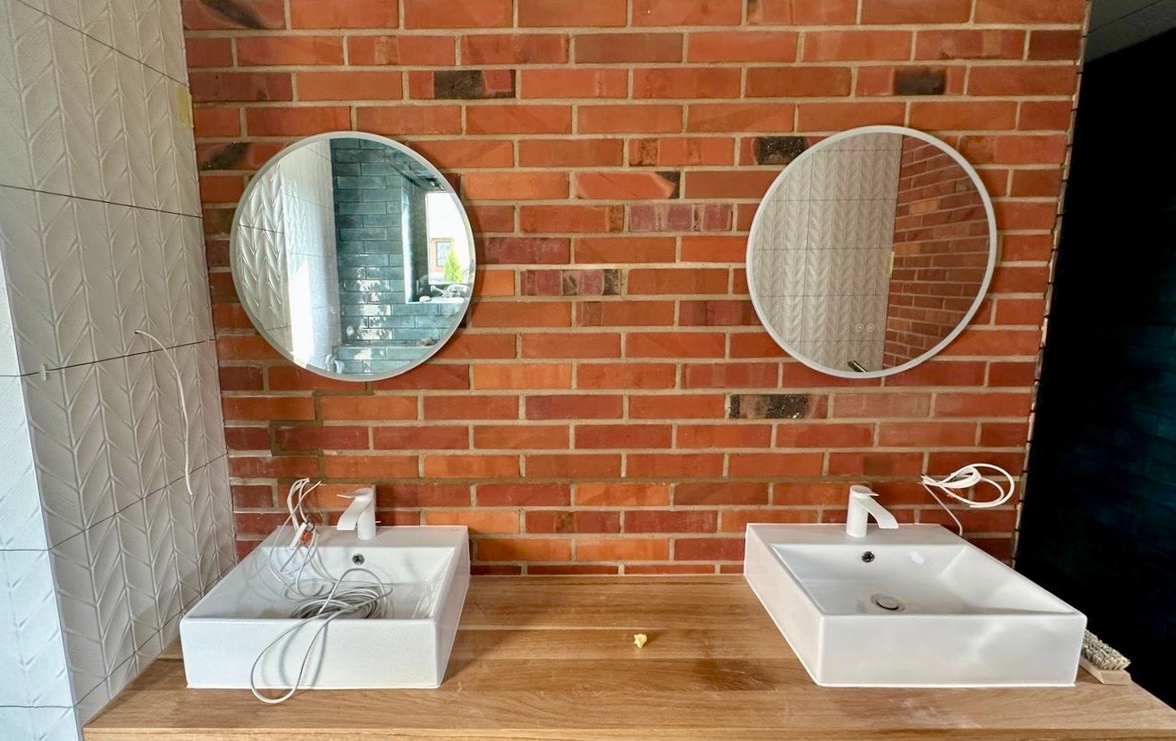 Dwie białe, prostokątne umywalki z bateriami na drewnianym blacie, zamontowane na tle ściany z czerwonej cegły i białych płytek 3D, z okrągłymi lustrami nad nimi, w trakcie montażu instalacji.