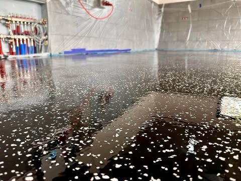 Błyszcząca, ciemna podłoga z białymi drobinkami, świeżo wylana, w pomieszczeniu zabezpieczonym folią malarską, widoczne rury grzewcze przy ścianie.