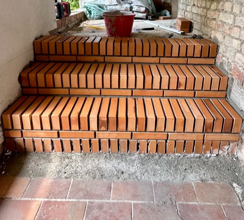 Schody wyłożone cegłą klinkierową w trakcie budowy, widoczne szare spoiny i czerwone wiadro na górnym stopniu.