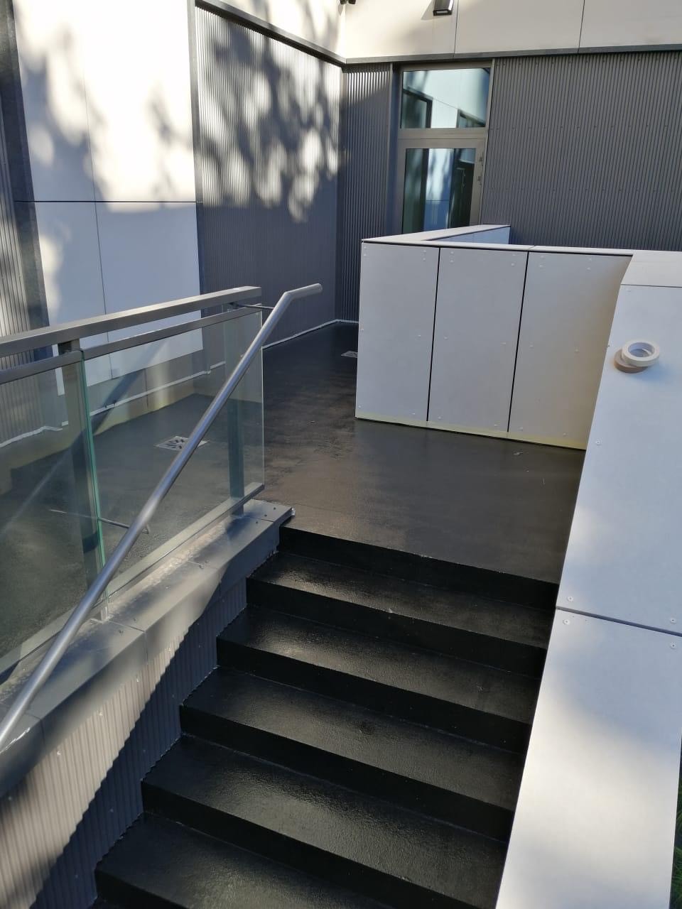 Schody pomalowane na czarno z metalową barierką i szklanym wypełnieniem, widoczne elementy elewacji budynku w odcieniach szarości i bieli, taśma malarska na krawędzi.
