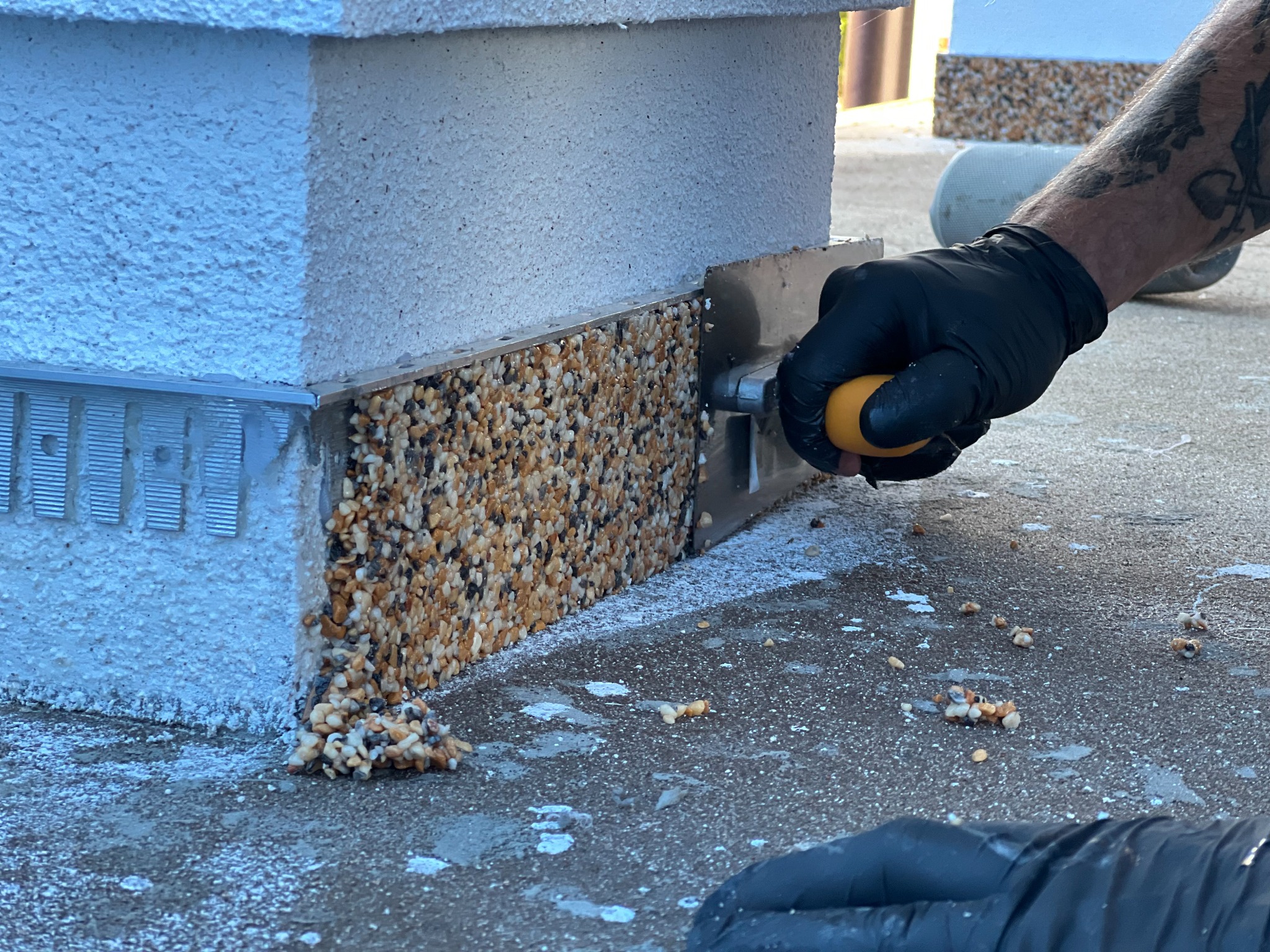 Nakładanie mozaiki z drobnych kamyczków na cokół budynku za pomocą pacy, widoczne pozostałości materiału na podłożu, ręka w czarnej rękawicy z tatuażem.