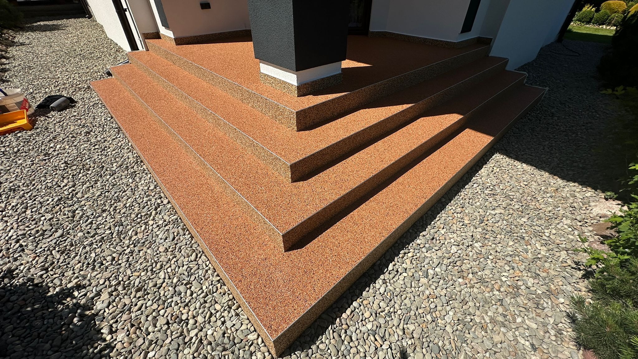 Zewnętrzne schody wejściowe z żywicznej, pomarańczowo-brązowej posadzki, otoczone dekoracyjnym żwirem, z widocznym fragmentem elewacji budynku i czarną kolumną.