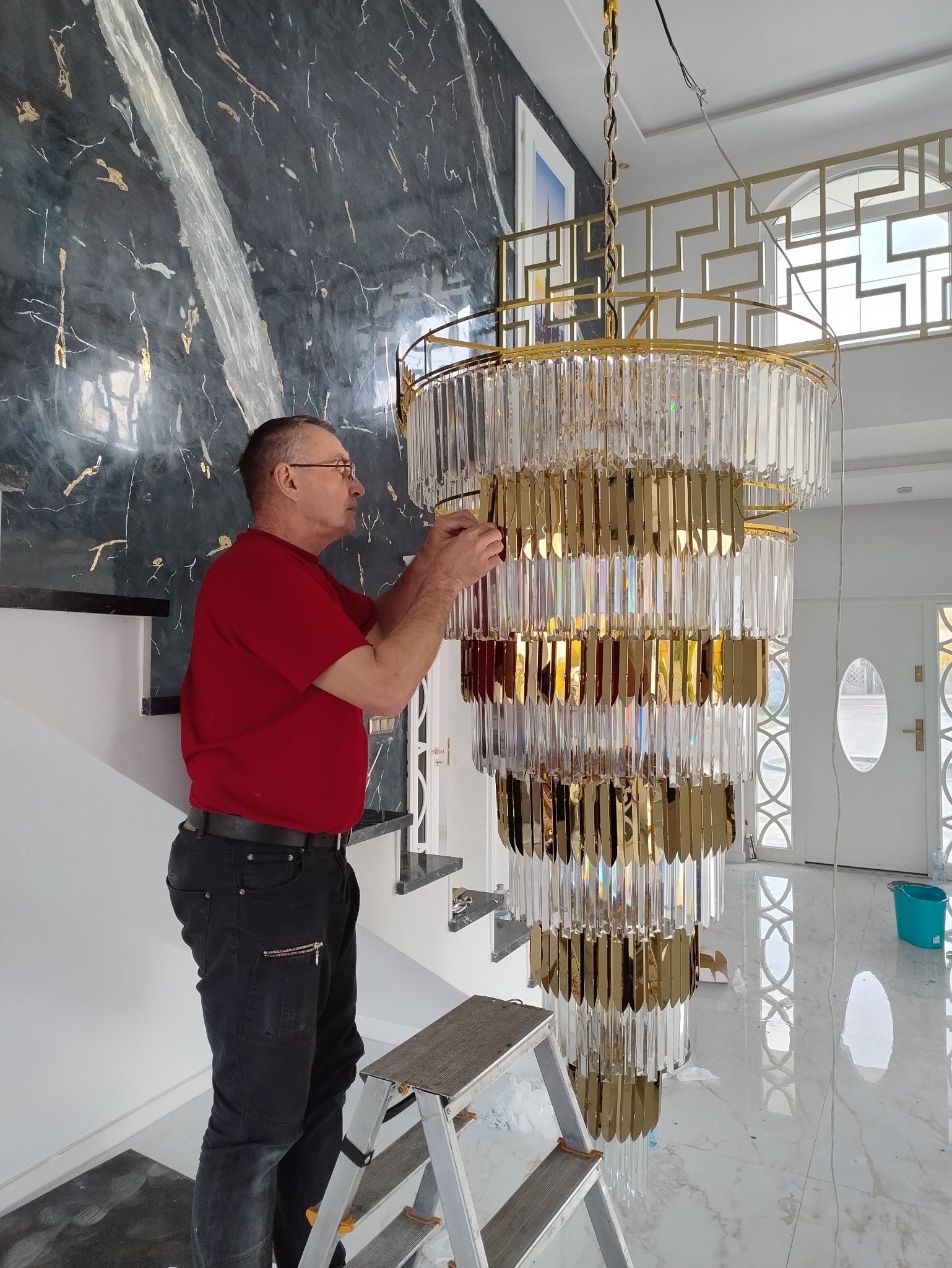 Elektryk na drabinie montuje duży, nowoczesny żyrandol kryształowo-złoty w przestronnym holu z marmurową posadzką i ścianą o wzorze marmuru.