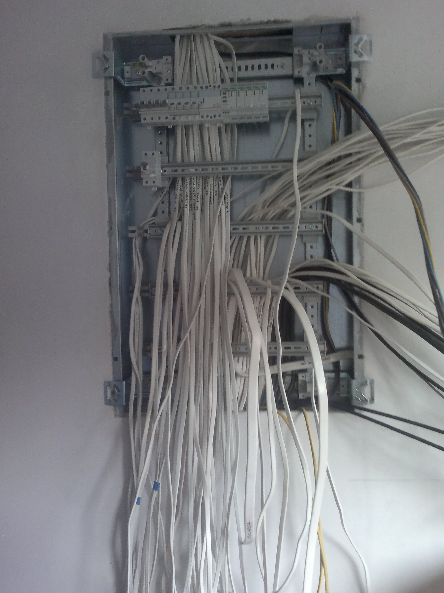 Otwarta skrzynka elektryczna w ścianie z licznymi przewodami elektrycznymi o różnych kolorach (białe, czarne, żółte) wychodzącymi z niej, widoczne elementy mocujące i listwy zaciskowe wewnątrz.