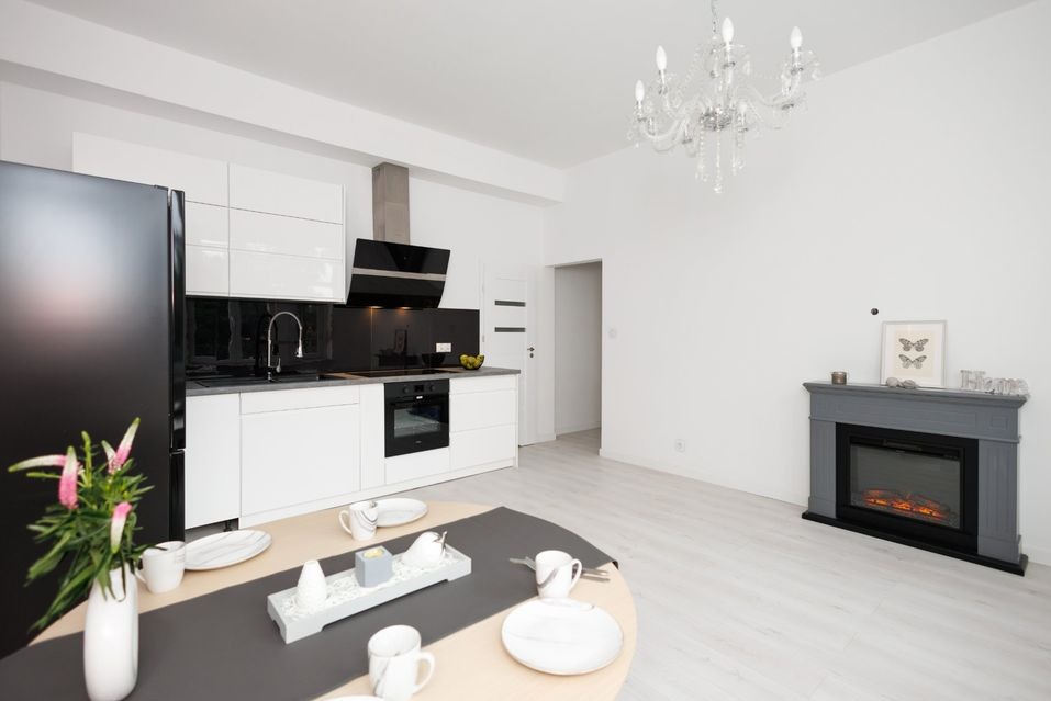 Nowoczesne wnętrze: biała kuchnia z czarnym akcentem, okrągły stół z zastawą, dekoracyjny kominek z szarym obramowaniem i kryształowy żyrandol, całość w jasnej tonacji.