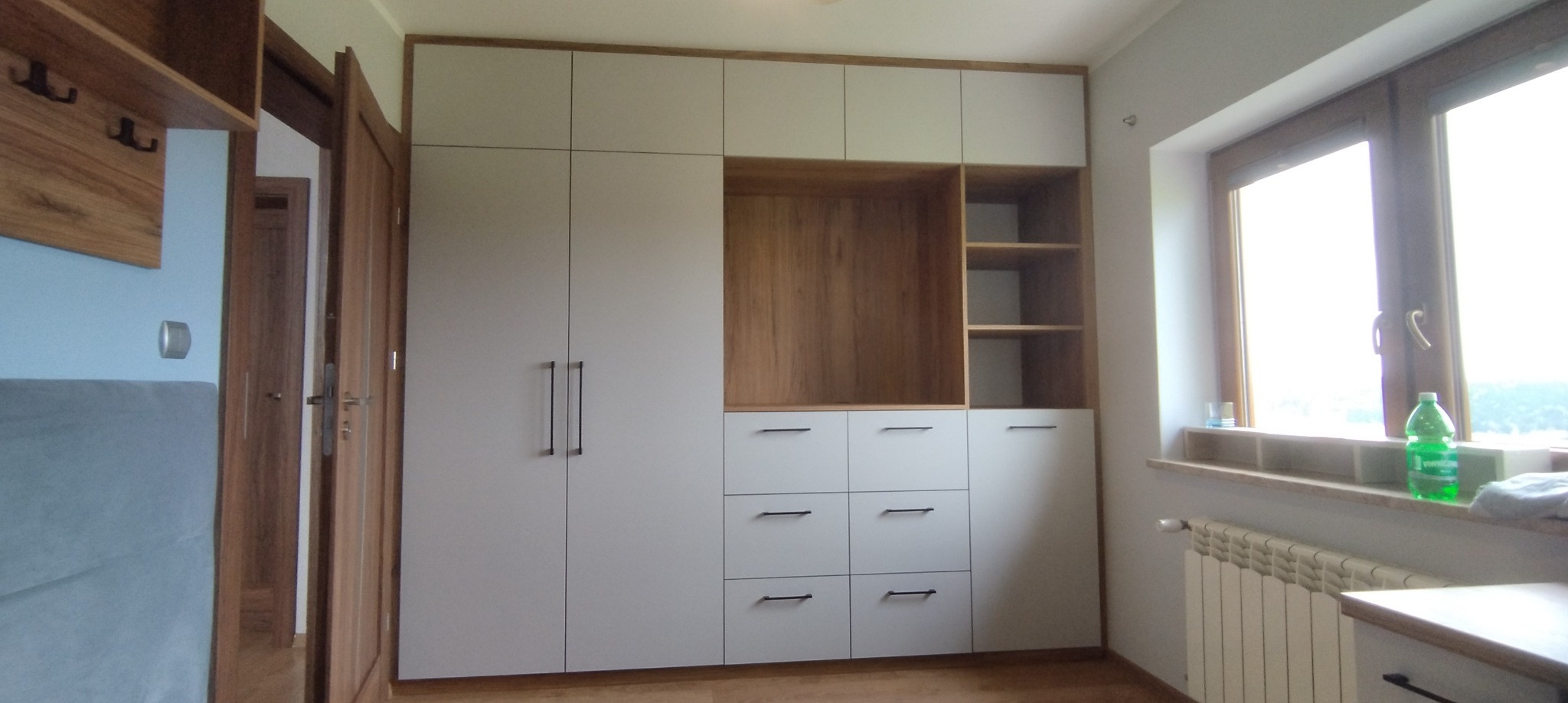 Biała szafa wnękowa z drewnianymi akcentami i czarnymi uchwytami, z otwartą półką i szufladami, stojąca przy oknie z butelką.