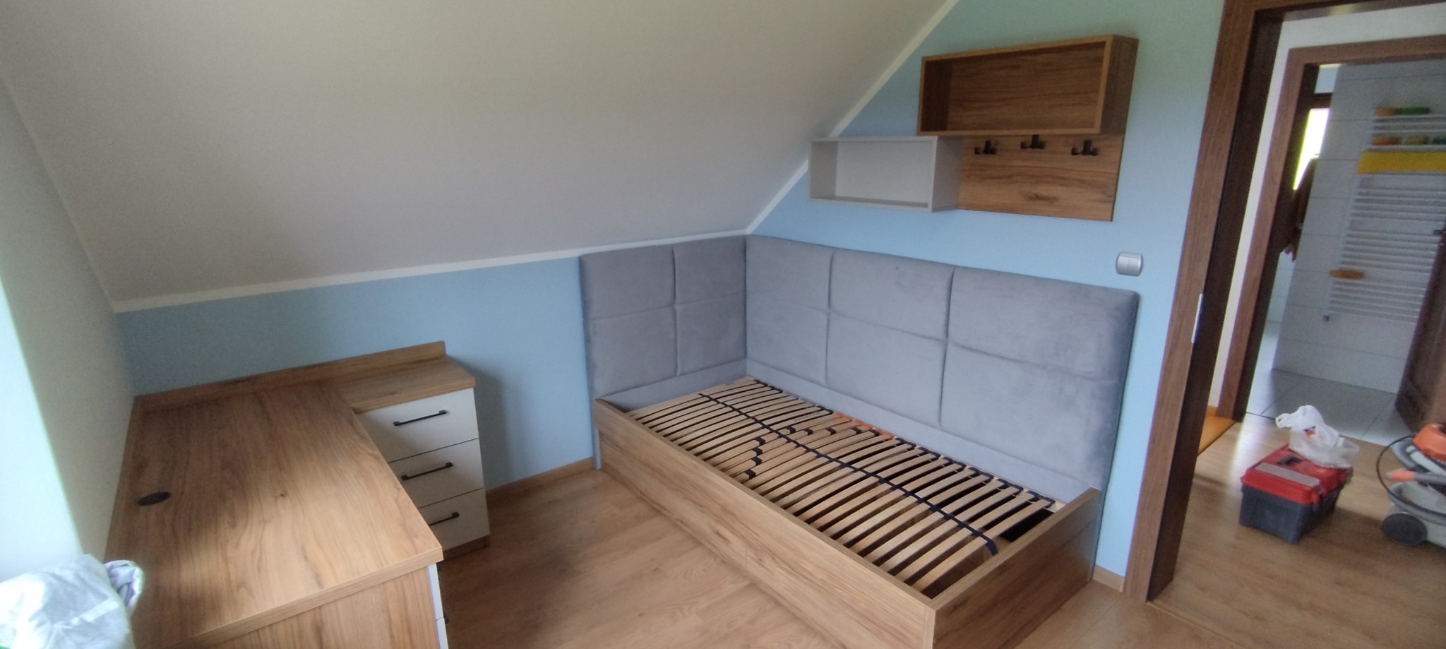 Pokój na poddaszu z drewnianą zabudową: biurko z szufladami, łóżko z tapicerowanym wezgłowiem i drewniane półki wiszące z wieszakami.