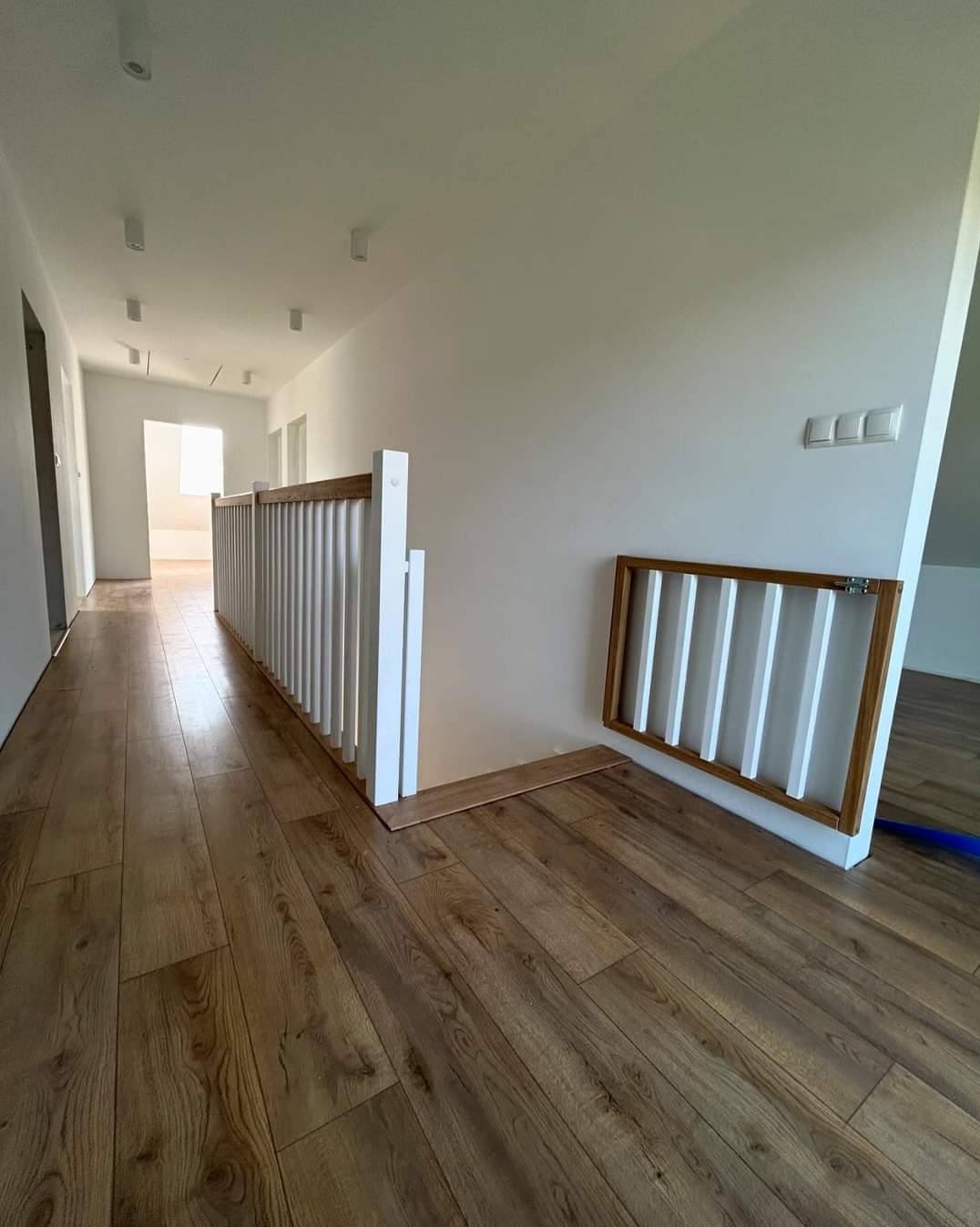 Jasny korytarz z drewnianą podłogą, białą balustradą schodową i bramką zabezpieczającą, widoczne panele podłogowe niedokończone przy balustradzie.