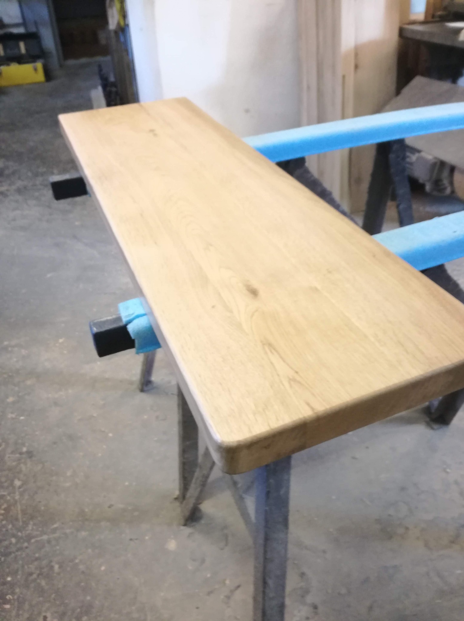Obrabiany drewniany blat na metalowych kozłach z zabezpieczeniem z niebieskiej pianki, widoczne ślady obróbki.
