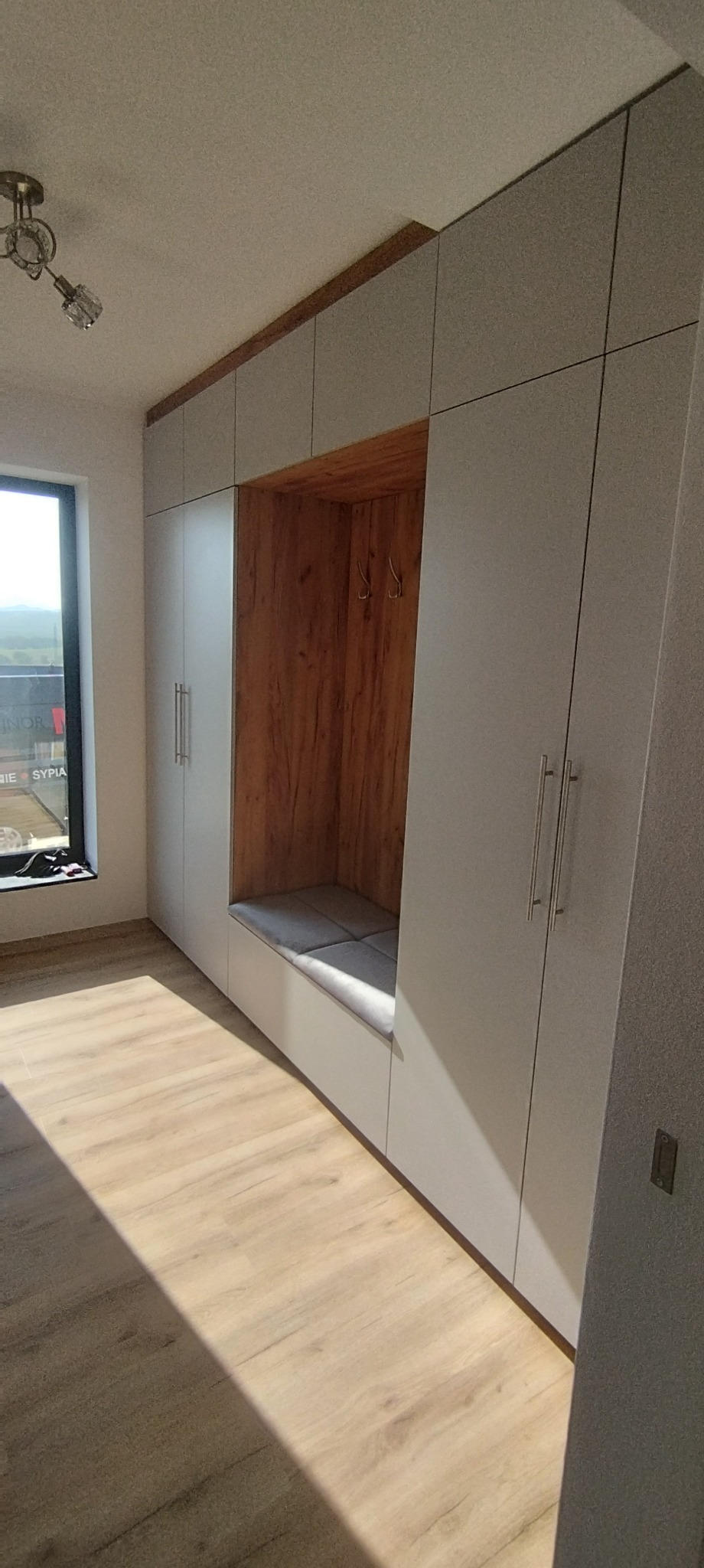 Szara szafa wnękowa z siedziskiem wykończonym drewnem, z widocznymi wieszakami wewnątrz, oświetlona naturalnym światłem z okna po lewej stronie.