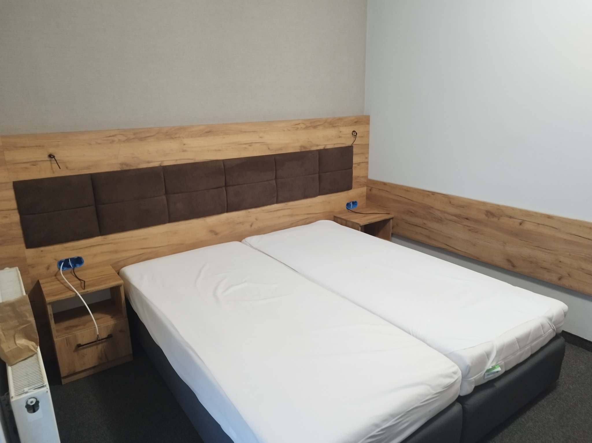 Dwa pojedyncze łóżka z białymi prześcieradłami, drewnianym wezgłowiem z brązowymi panelami tapicerowanymi, niedokończone podłączenie elektryczne obok szafki nocnej.