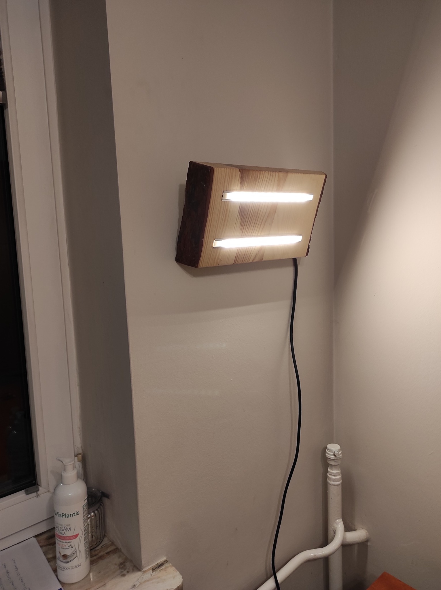 Kinkiet drewniany wykonany z fragmentu pnia z dwoma wbudowanymi świetlówkami LED, przymocowany do ściany, z widocznym kablem zasilającym.