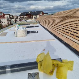 F.h.u. Adam Danczak - Montaż drewnianej konstrukcji dachu na białej membranie, widoczne rolki papy, worki z materiałem i fragmenty osiedla w tle.