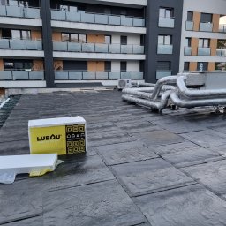 F.h.u. Adam Danczak - Płaski dach budynku wielorodzinnego z papy bitumicznej, widoczne rury instalacyjne w srebrnej izolacji oraz paczka styropianu do ocieplenia.