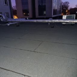 F.h.u. Adam Danczak - Płaski dach pokryty papą z widoczną instalacją wentylacyjną, rurami w srebrnej izolacji oraz klimatyzatorem na tle budynków miejskich o zmierzchu.