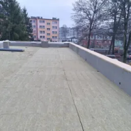 Płaski dach w trakcie ocieplania, widoczne płyty izolacyjne, obrzeża z betonu i drewna, w tle budynki mieszkalne i drzewa.