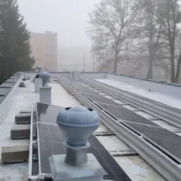 Płaski dach pokryty jasną membraną z zainstalowanymi panelami fotowoltaicznymi i szarymi kominkami wentylacyjnymi na tle zamglonego krajobrazu miejskiego z drzewami i budynkiem.