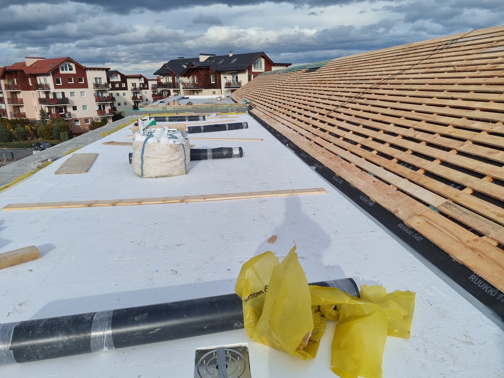 Montaż drewnianej konstrukcji dachu na białej membranie, widoczne rolki papy, worki z materiałem i fragmenty osiedla w tle.