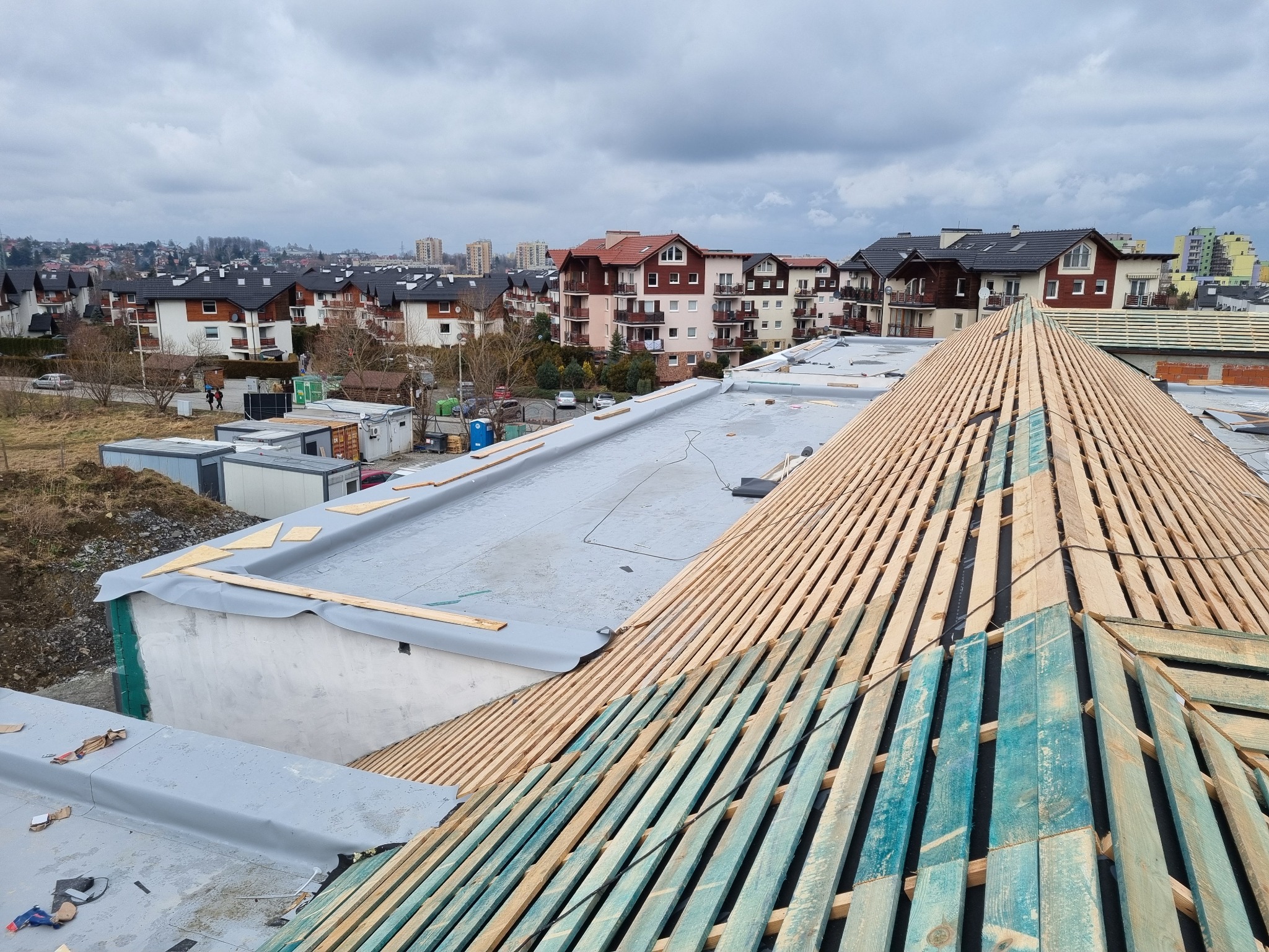 Konstrukcja więźby dachowej z desek impregnowanych na niebiesko, widok z góry na tle zabudowy Domaniewic i pochmurnego nieba.