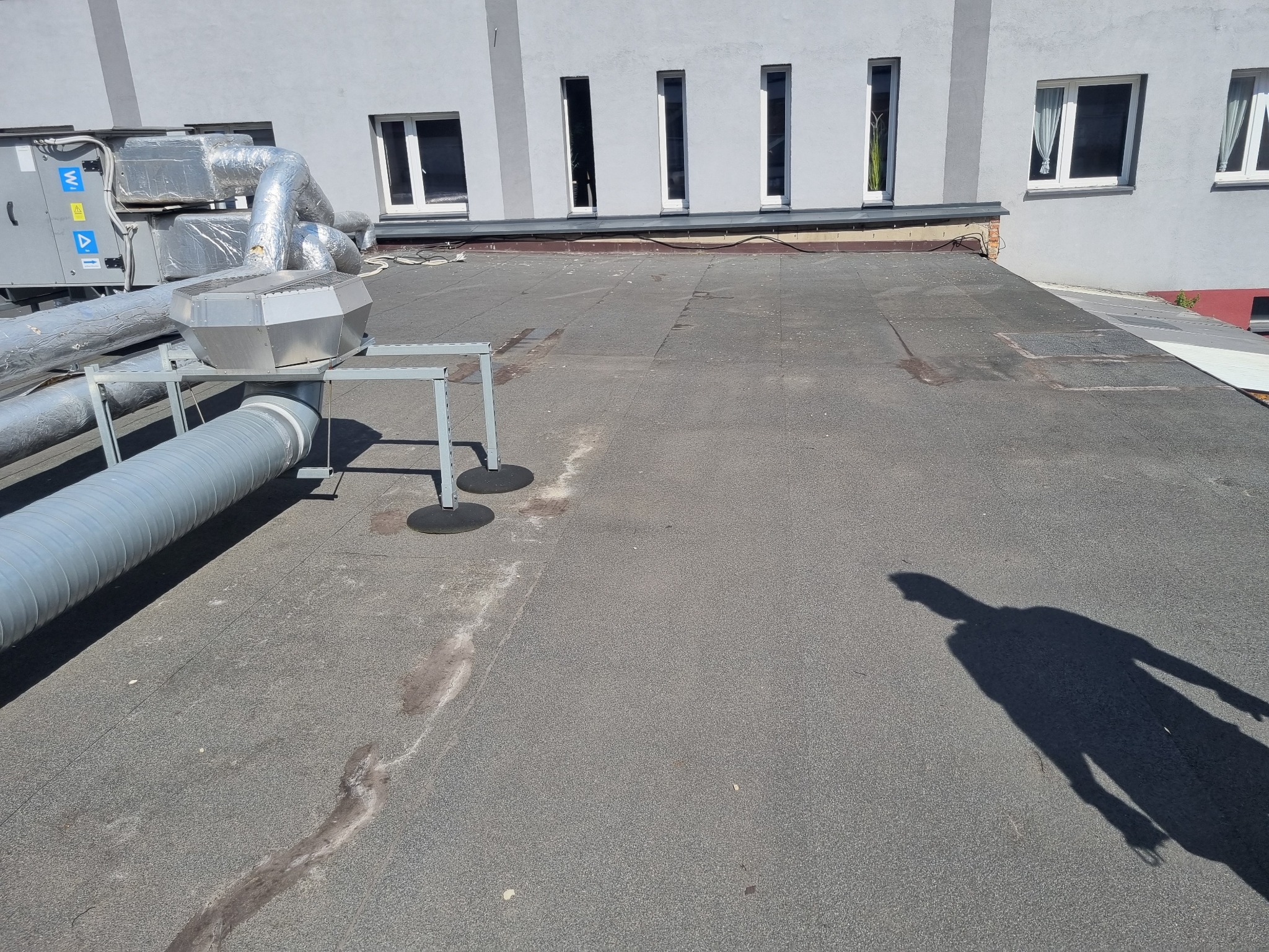 Płaski dach pokryty papą z widocznymi instalacjami wentylacyjnymi i klimatyzacyjnymi, cień osoby na powierzchni dachu, szary budynek w tle z rzędami okien.