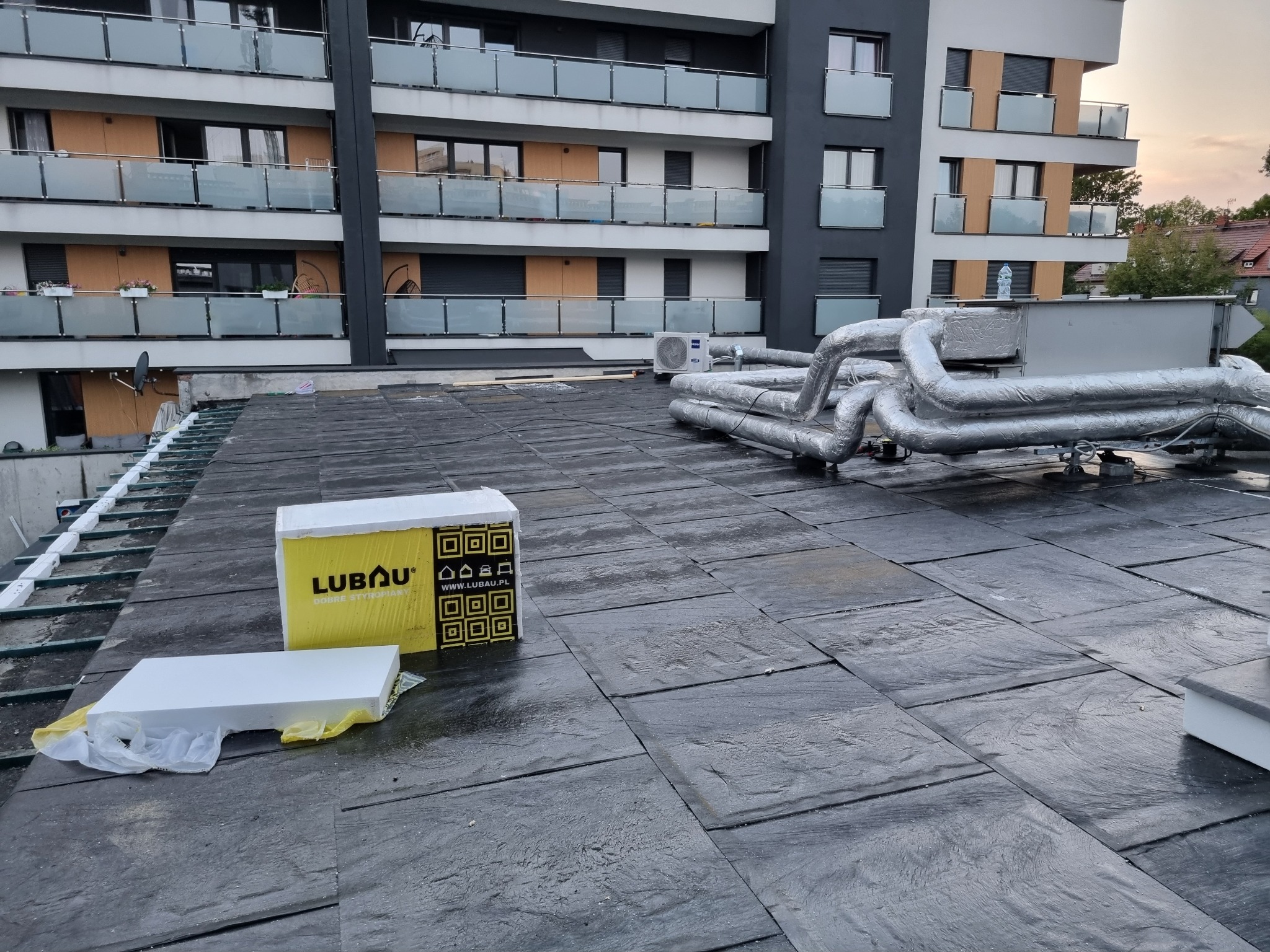 Płaski dach budynku wielorodzinnego z papy bitumicznej, widoczne rury instalacyjne w srebrnej izolacji oraz paczka styropianu do ocieplenia.