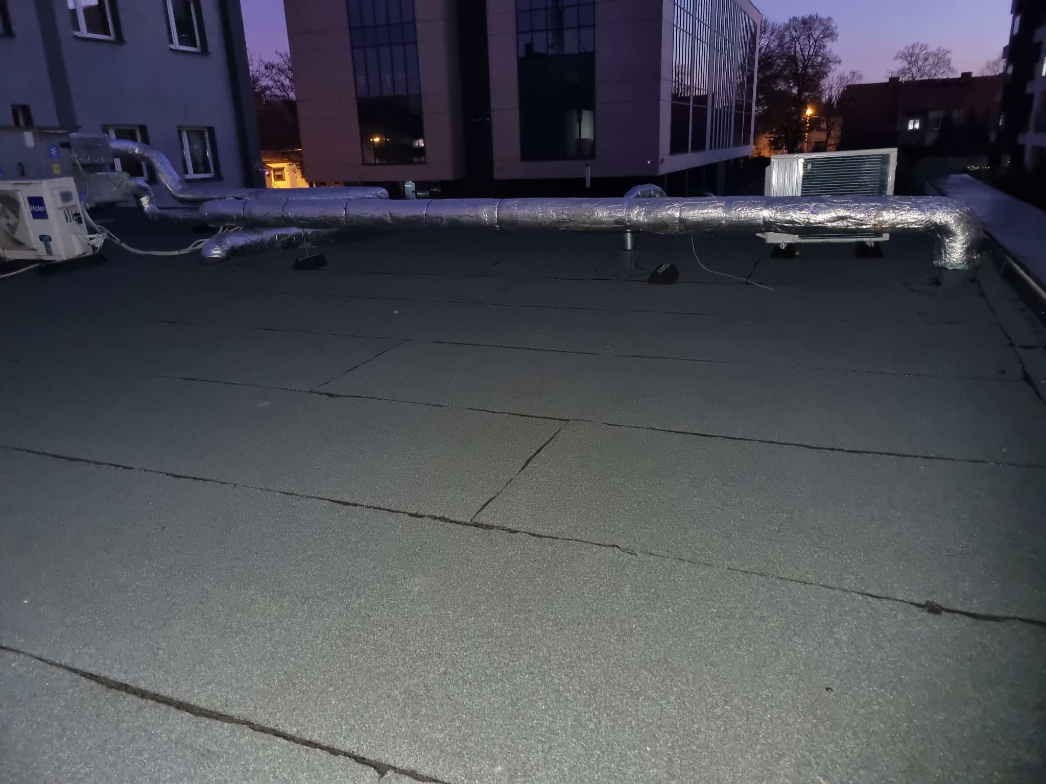 Płaski dach pokryty papą z widoczną instalacją wentylacyjną, rurami w srebrnej izolacji oraz klimatyzatorem na tle budynków miejskich o zmierzchu.