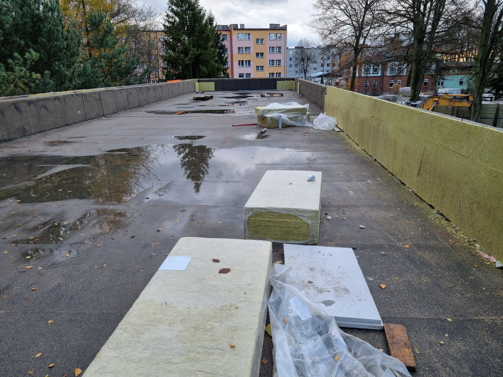 Płaski dach pokryty papą, częściowo mokry po deszczu, z rozłożonymi paczkami wełny mineralnej i płytami styropianowymi przygotowanymi do ocieplenia, na tle bloków mieszkalnych i drzew