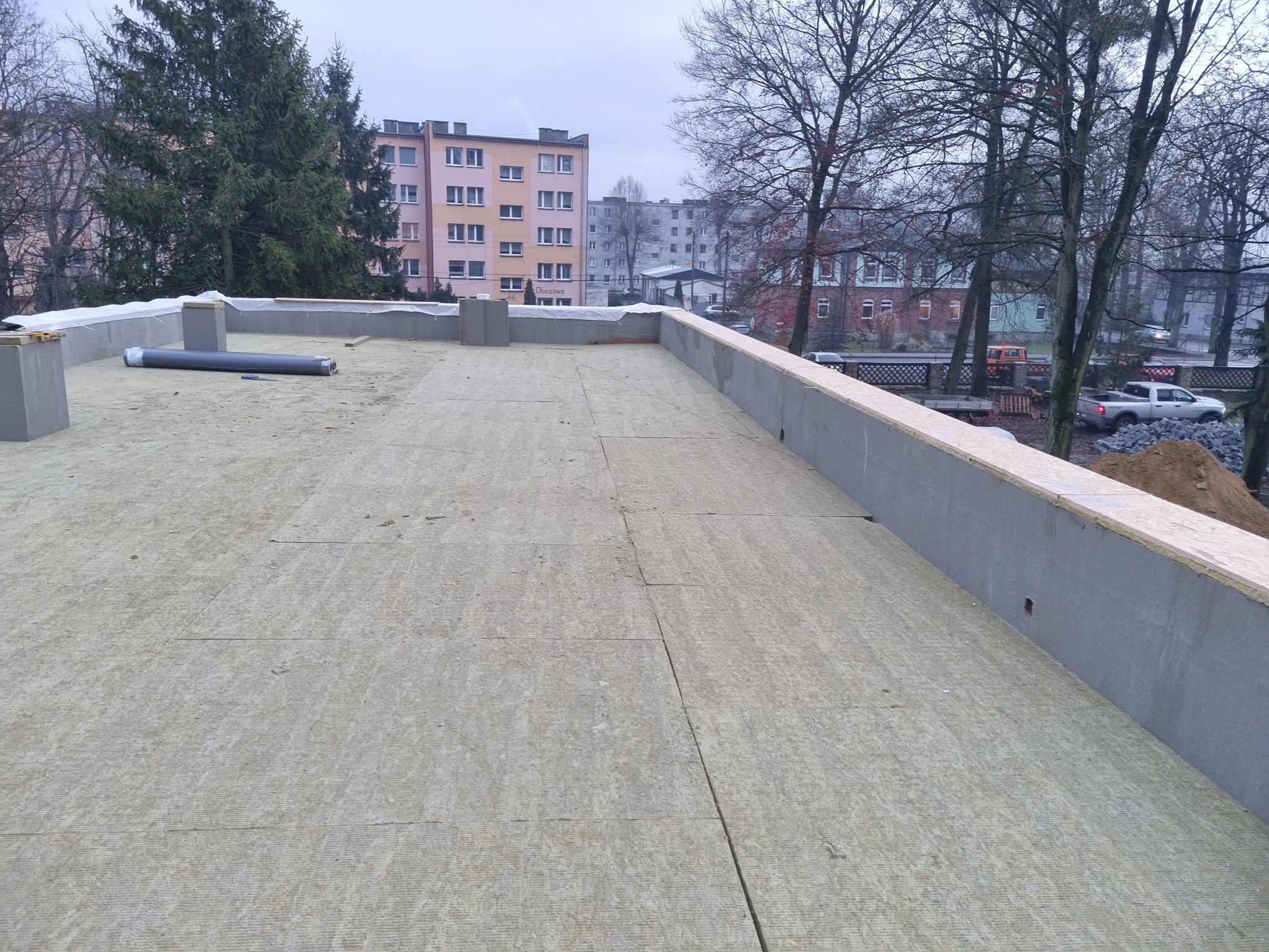 Płaski dach w trakcie ocieplania, widoczne płyty izolacyjne, obrzeża z betonu i drewna, w tle budynki mieszkalne i drzewa.