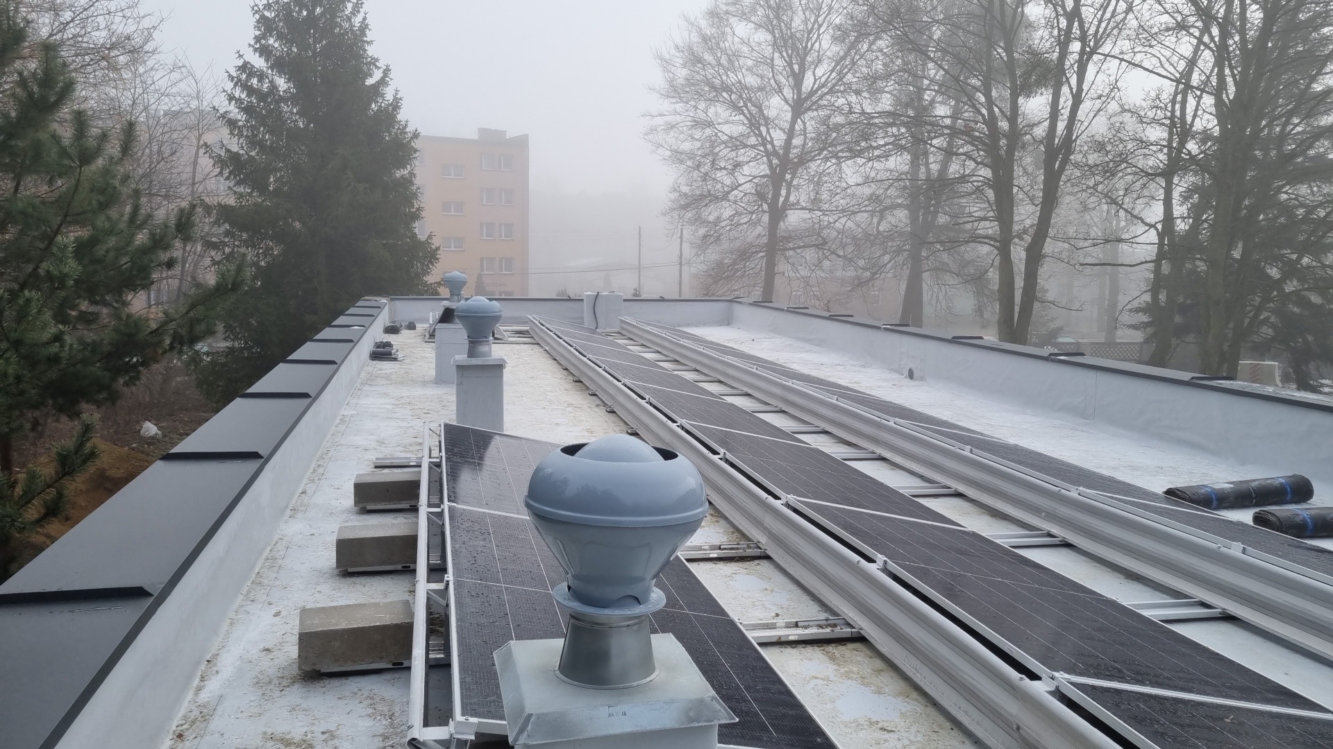 Płaski dach pokryty jasną membraną z zainstalowanymi panelami fotowoltaicznymi i szarymi kominkami wentylacyjnymi na tle zamglonego krajobrazu miejskiego z drzewami i budynkiem.