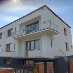 R-BUD - Dwukondygnacyjny dom z białymi ścianami i ciemną podmurówką, balkony z białą balustradą, brązowe ramy okienne, widok z dołu, słoneczny dzień.