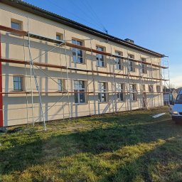 R-BUD - Budynek w trakcie prac elewacyjnych, otoczony rusztowaniem. Na pierwszym planie trawnik i zaparkowany biały bus. Słoneczny dzień.