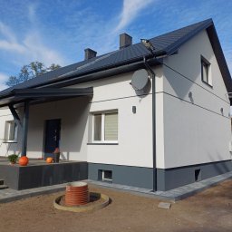 R-BUD - Nowoczesny dom z jasną elewacją i ciemnoszarą podmurówką, widoczne schody wejściowe i elementy dekoracyjne. Dach dwuspadowy z ciemnej dachówki. Błękitne niebo w tle.