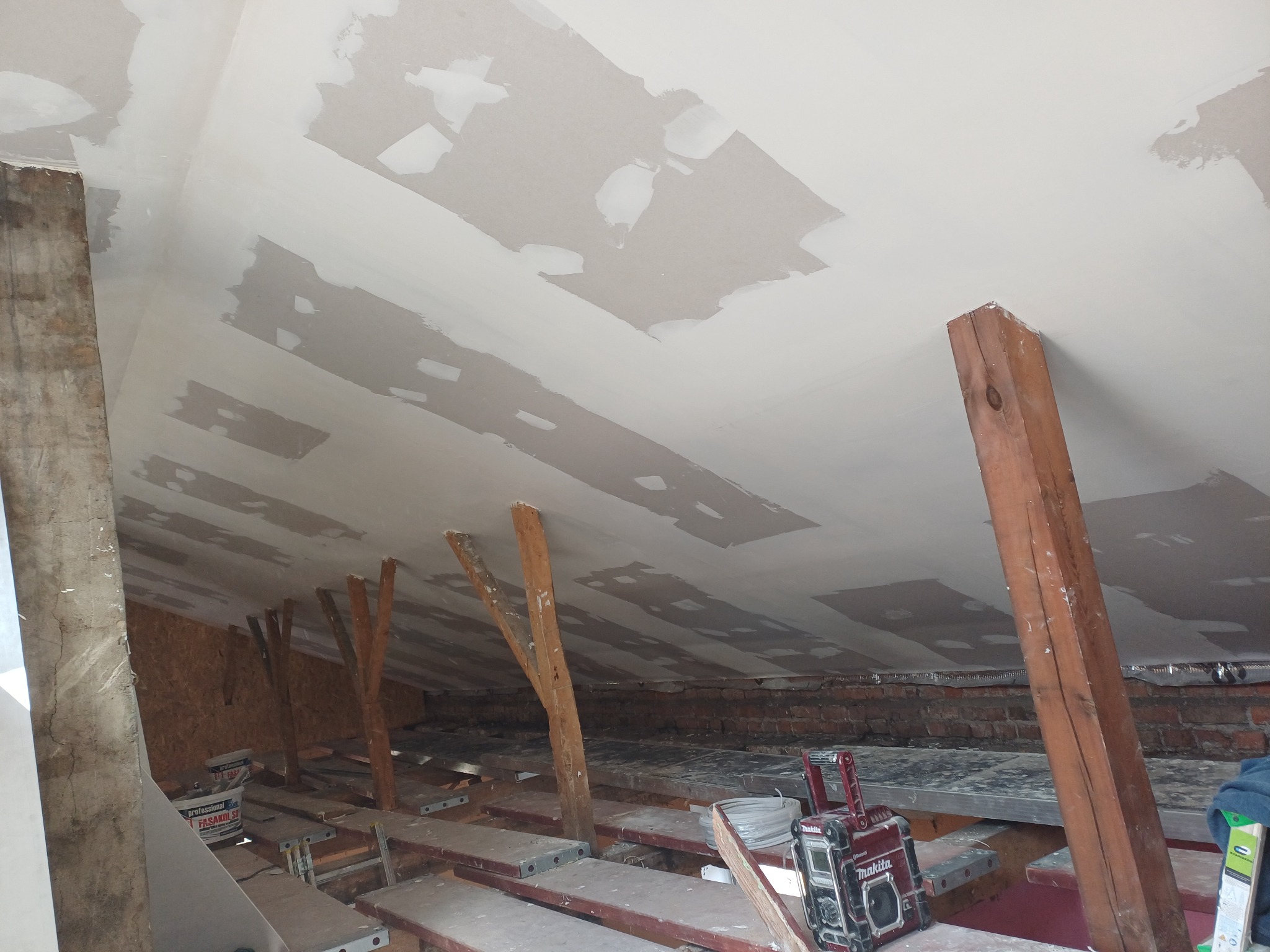 Skośny sufit poddasza w trakcie prac wykończeniowych, widoczne łaty gipsowe, drewniane belki konstrukcyjne i narzędzia. Ujęcie z dołu, z bliska.