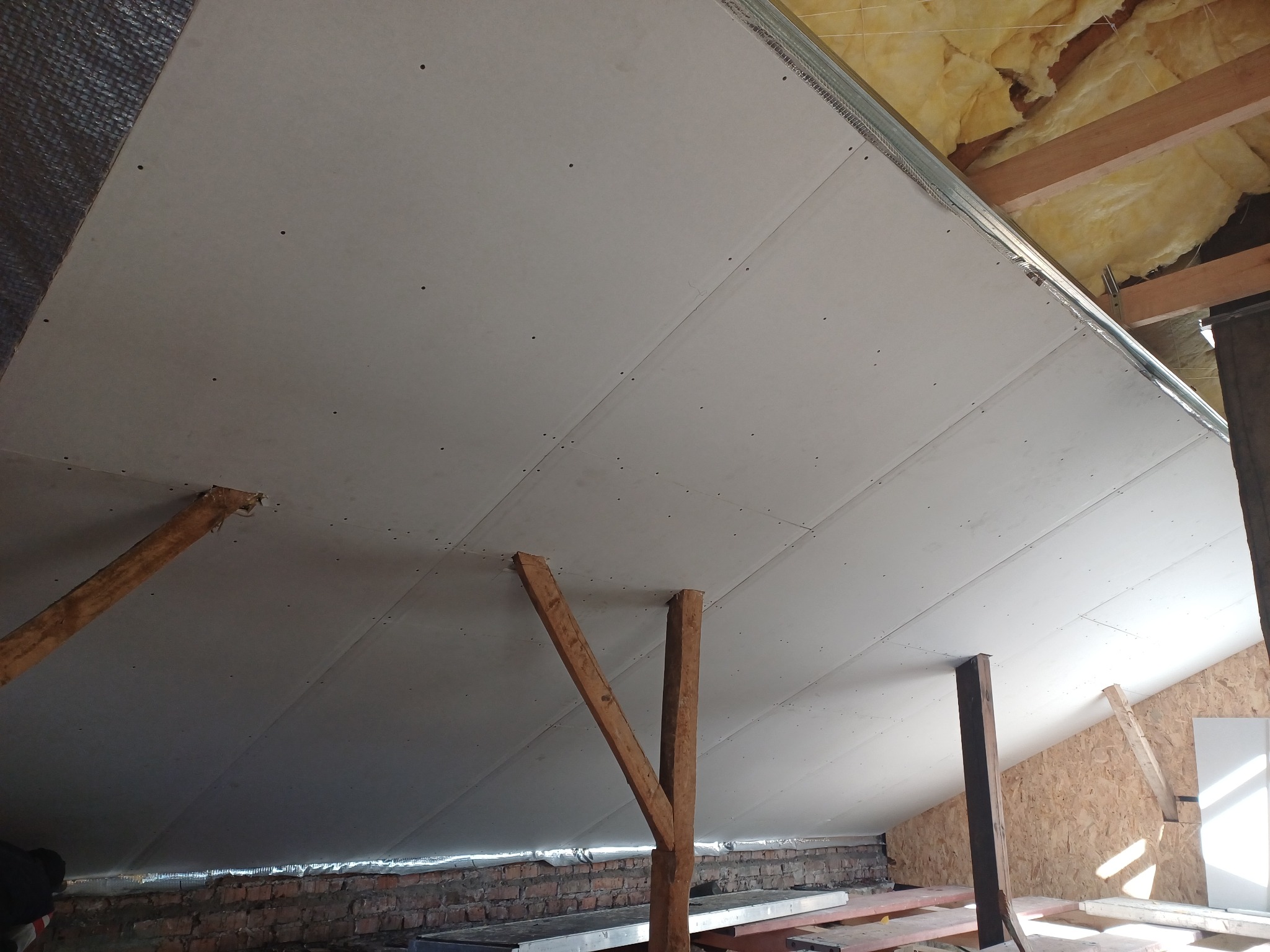 Płyty gipsowo-kartonowe na poddaszu, widoczne drewniane podpory i ocieplenie. Etap prac wykończeniowych, sufit pod skosem.