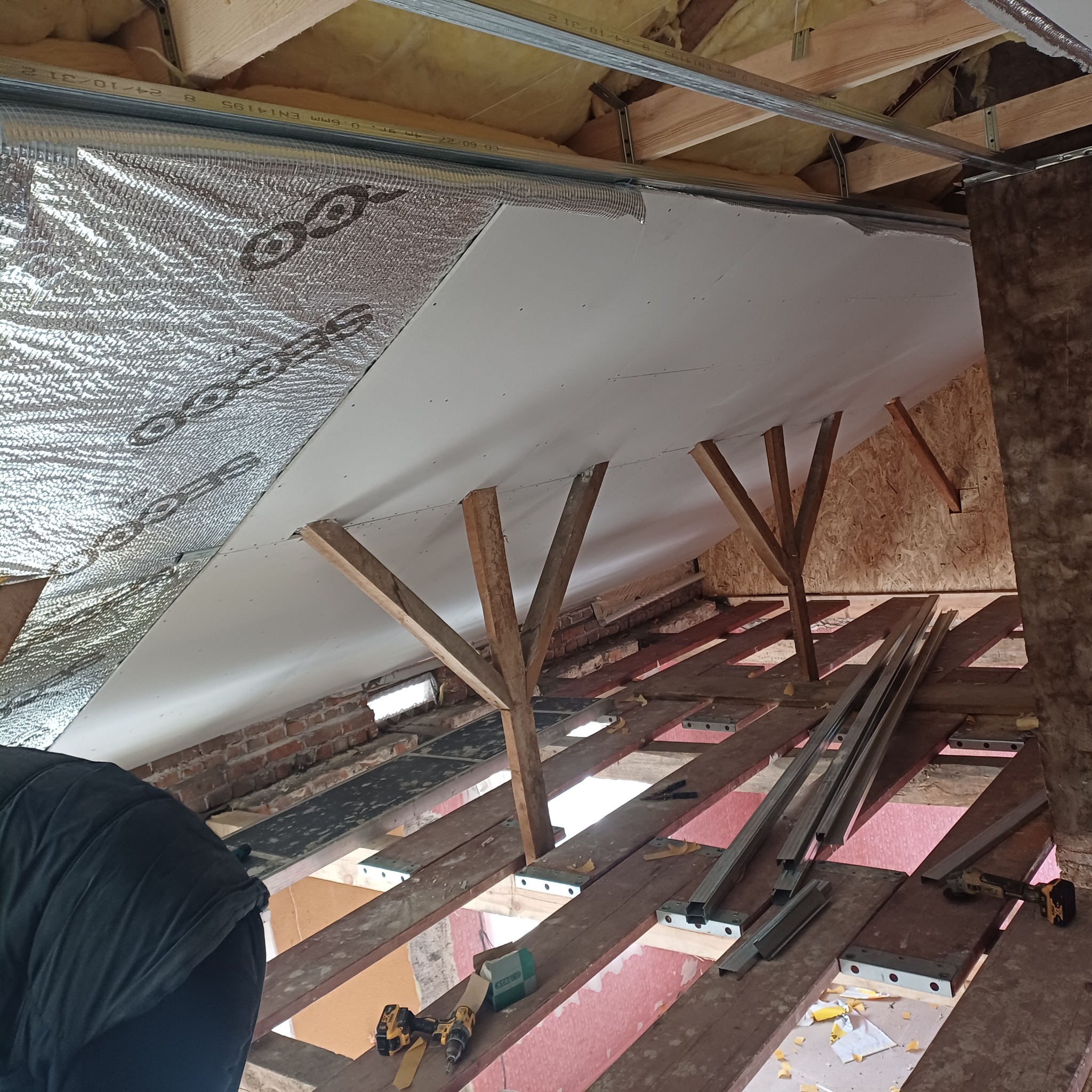 Montaż izolacji termicznej i płyt gipsowo-kartonowych na poddaszu. Widoczne drewniane podpory, folia aluminiowa i narzędzia. Prace wykończeniowe w trakcie realizacji.