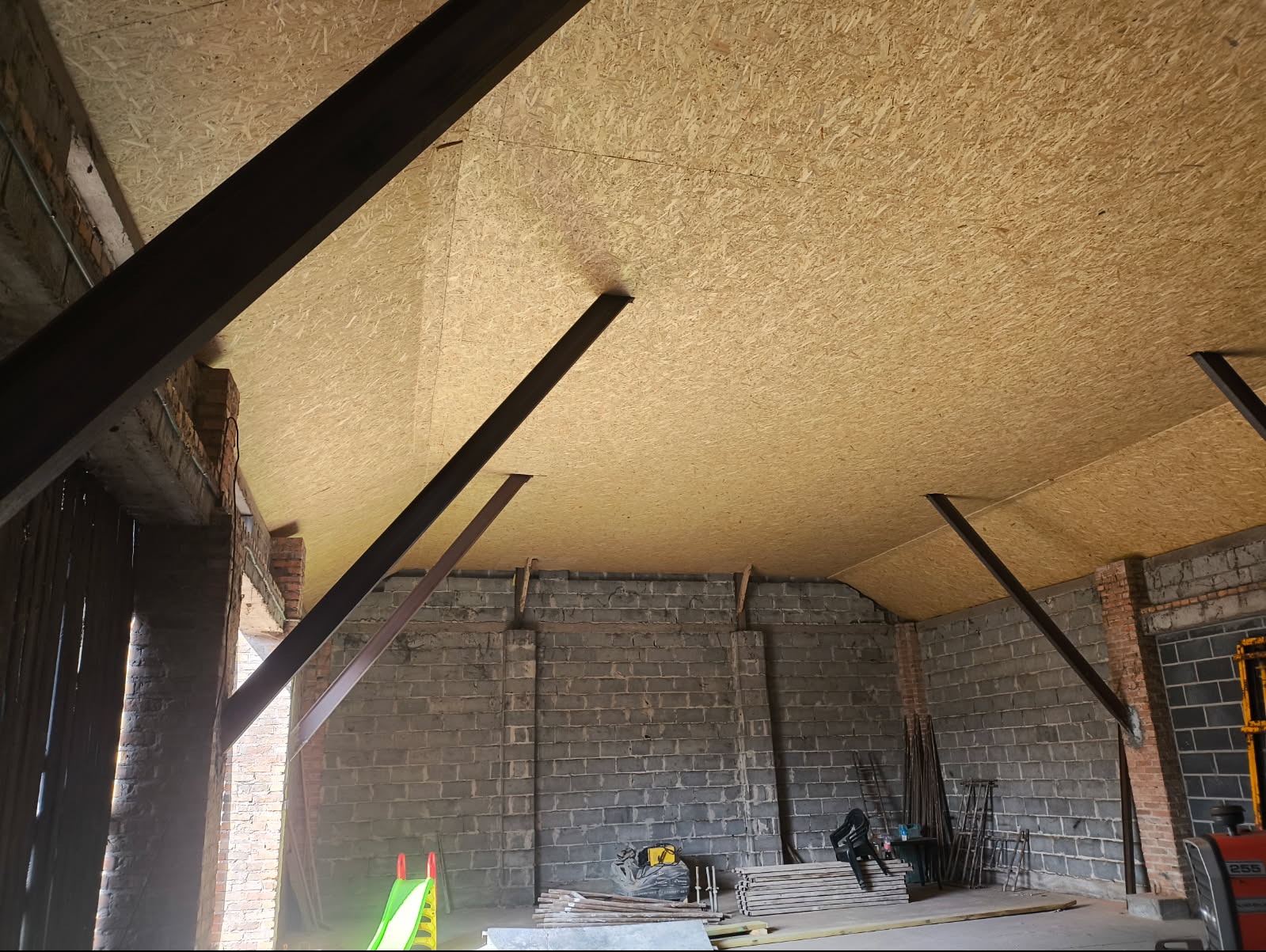 Płyta OSB na suficie poddasza, wsparta stalowymi belkami. Surowy mur z cegły i betonu w tle. Widoczne narzędzia i materiały budowlane. Etap prac wykończeniowych.