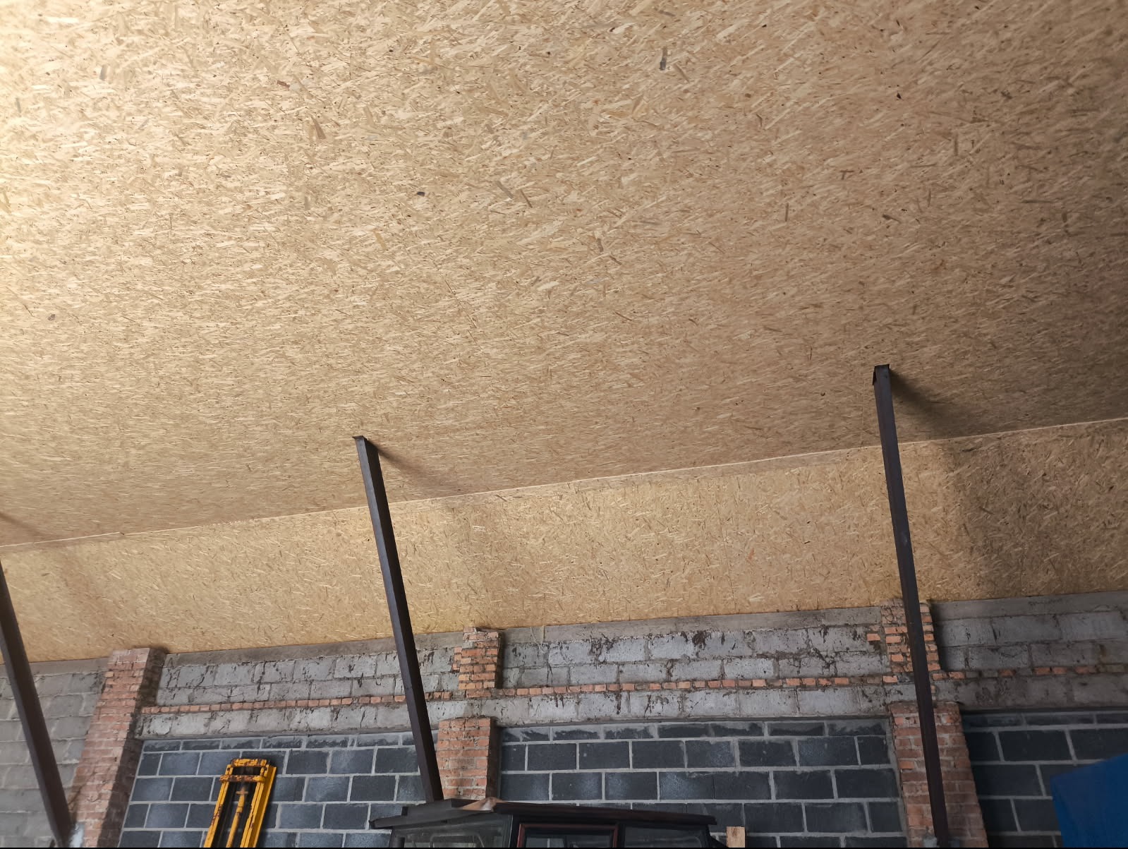 Płyta OSB na suficie, wsparta metalowymi słupkami, widok z dołu. Fragment murowanej ściany z ceglanymi elementami w tle. Prace wykończeniowe wewnątrz budynku.
