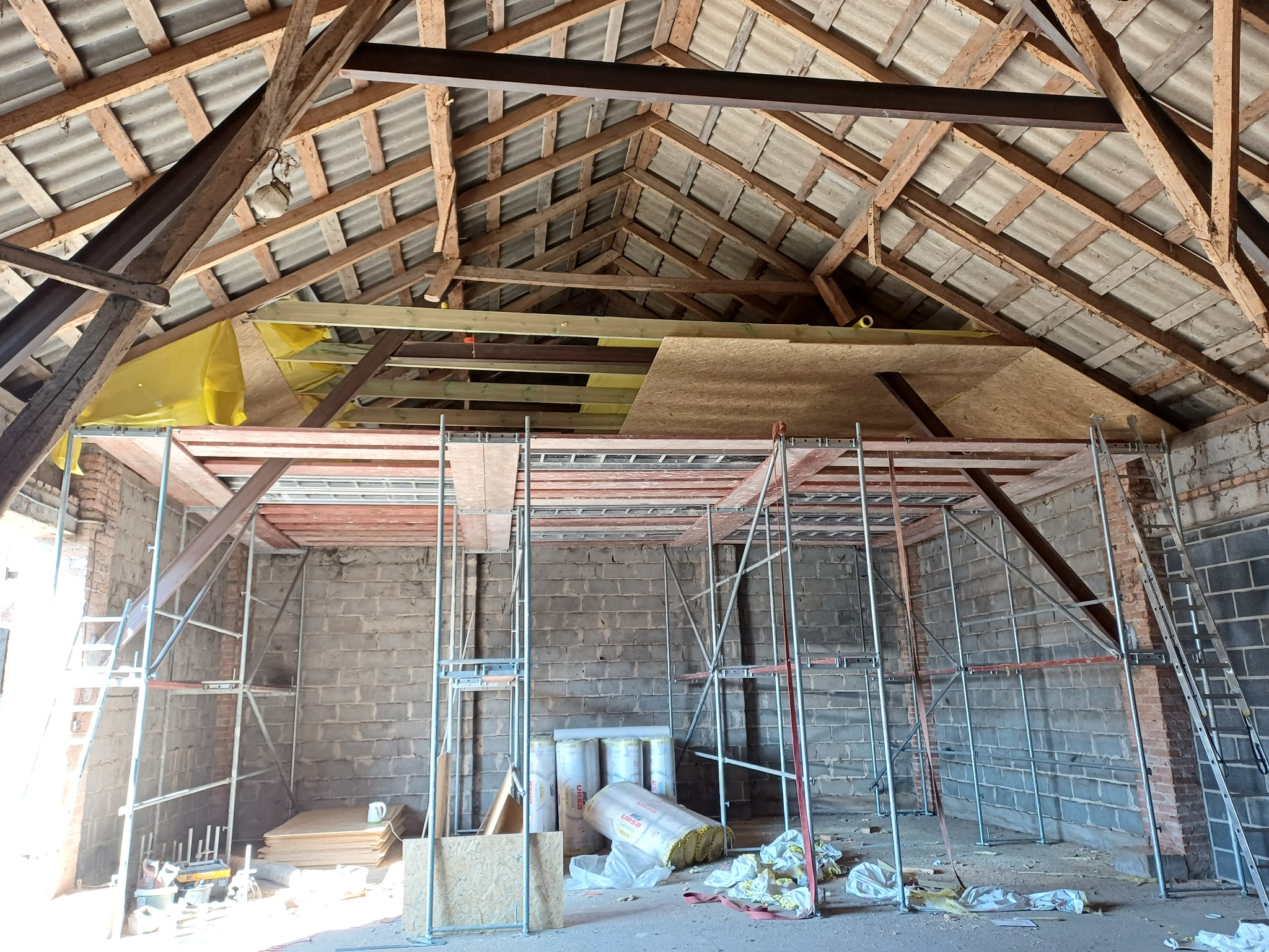 Szkielet dachu z widocznymi elementami konstrukcyjnymi i rusztowaniem. W tle materiały izolacyjne, płyty OSB i narzędzia. Etap prac przygotowawczych do ocieplenia poddasza.