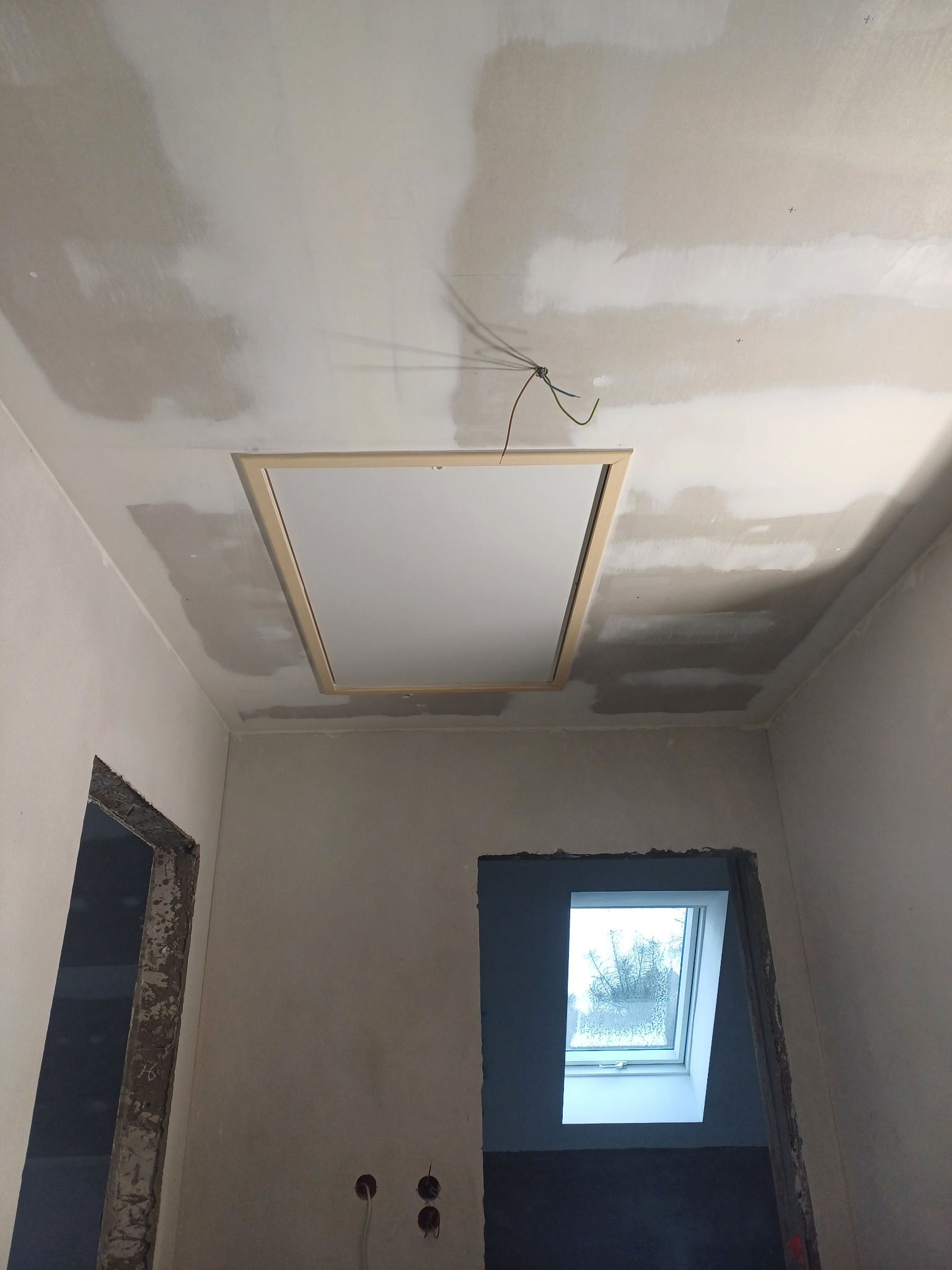 Wnętrze w trakcie prac wykończeniowych: sufit z zamontowanym włazem strychowym, ściany po nałożeniu gładzi, widoczne otwory okienne i drzwiowe w stanie surowym.