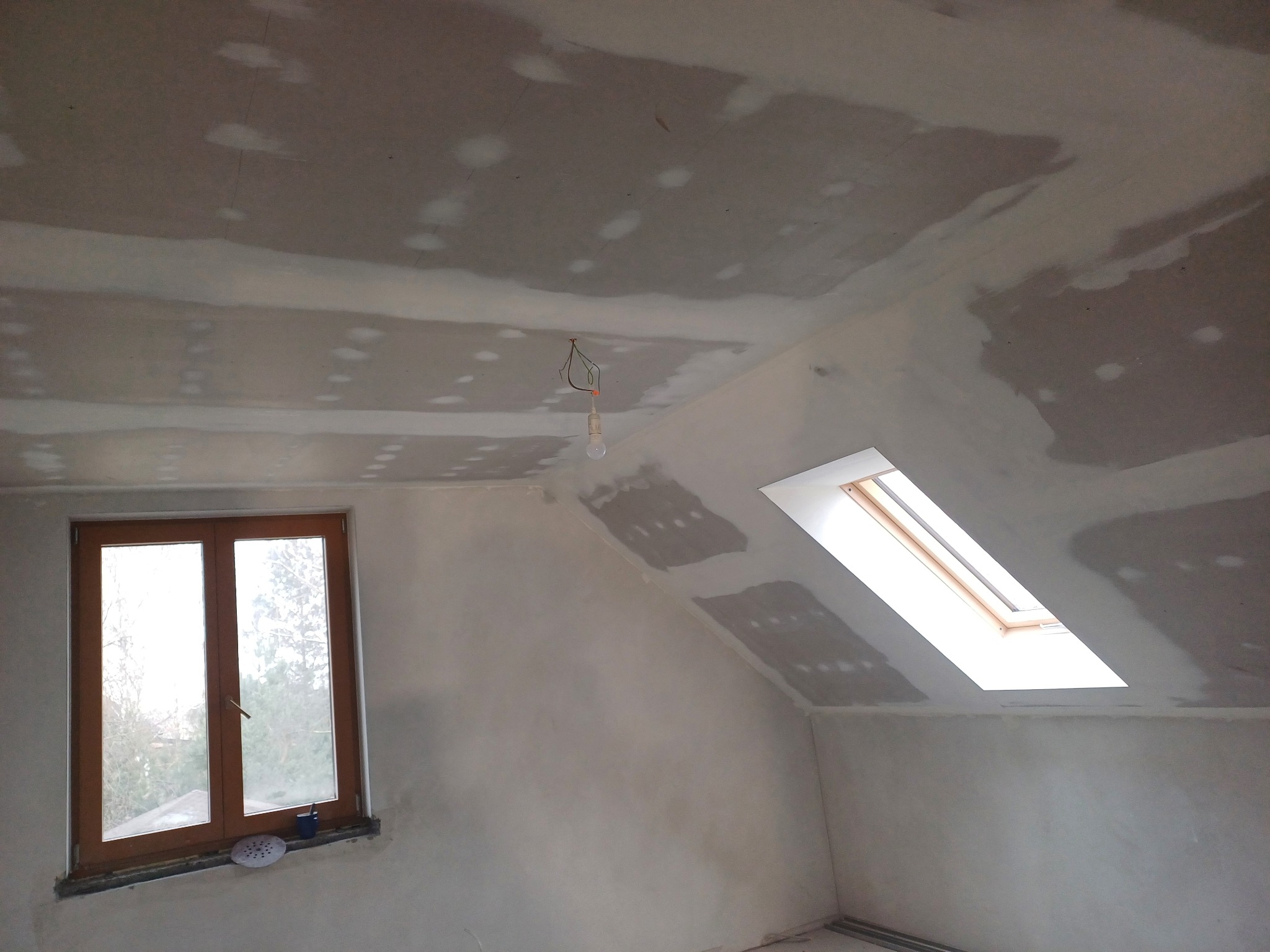 Poddasze ze ścianami i sufitem w trakcie szpachlowania. Widoczne okno połaciowe i zwykłe okno. W tle drzewa. Wystający kabel z żarówką.