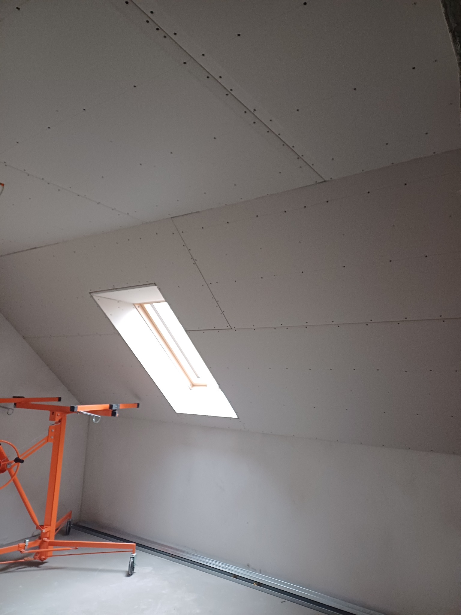 Poddasze z oknem dachowym w trakcie montażu płyt gipsowo-kartonowych, widoczny podnośnik do płyt. Szpachlowane łączenia płyt.